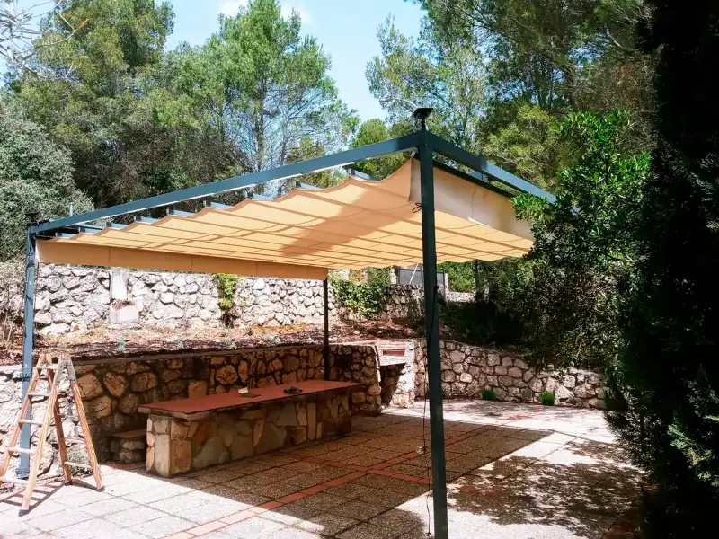 Toldo romano de lona clara sobre una pérgola junto a un quincho de piedra